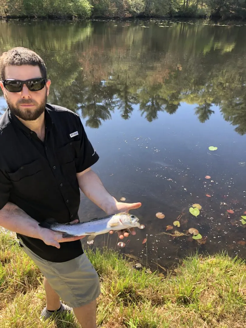 A Man With Black Shades Holding a Fish Infront of a Pond